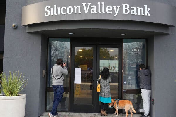 Three people outside a gray building with the words "Silicon Valley Bank" on it. Two people are looking at a sign posted on the building's glass doors. One person is taking photographs. 